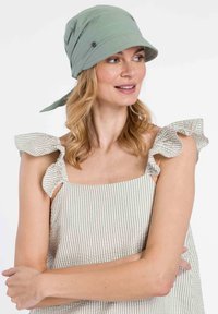Pălărie de tip bucket dintr-un material moale, verde deschis, cu un șnur, având un aspect subtil lucios. Modelul poartă un top fără mâneci, cu dungi, având accente volănoase la umeri.