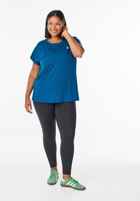 Modrá krátkastrihová blúzka s okrúhlym výstrihom, spojená s čiernymi legínami a zelenými teniskami. Tkanina sa zdá byť hladká a elastická.