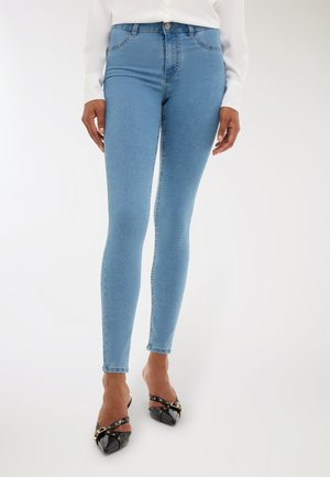 Donna che indossa jeans slim-fit azzurri chiari, un top bianco a maniche lunghe e décolleté neri con punta a stiletto e dettagli in oro.