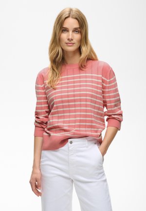 Femme aux longs cheveux blonds portant un pull rayé rose et blanc et un pantalon blanc, debout les mains dans les poches devant un fond uni.