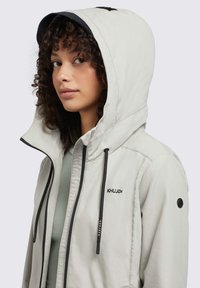Lichtgrijze ritsjack met capuchon, voorzien van zwarte trekkoorden en logo. Gladde textuur, schuine zakken en zwarte accenten op de capuchon.