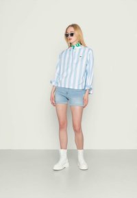 Camisa de gola riscada em azul claro e branco, calções de ganga e botins brancos de tornozelo. Um detalhe verde espreita do colarinho da camisa.
