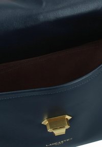 Borsa in pelle blu navy con una texture liscia, dotata di una chiusura in tono oro e un interno marrone. Include il logo del marchio impresso sul davanti.
