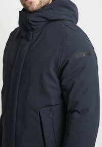 Marineblauwe hooded jack met een gladde, gestructureerde stof, zijzak met rits en een merkembleem op de schouder.