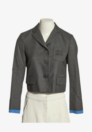 Blazer gris recortado con dos botones, puños azules, bolsillo en el pecho con bordado y pantalones blancos de cintura alta en un maniquí.