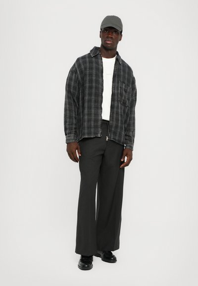 Camicia flanella a quadri in nero e grigio, T-shirt bianca sotto, pantaloni scuri dal taglio comodo e cappellino grigio. Scarpe nere completano il look.
