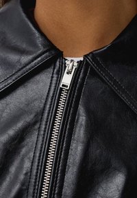 Czarna kurtka z sztucznej skóry z szerokim kołnierzem, z dodatkowym srebrnym zamkiem błyskawicznym i przeszyciami wzdłuż brzegów. Gładka tekstura i dopasowany krój.