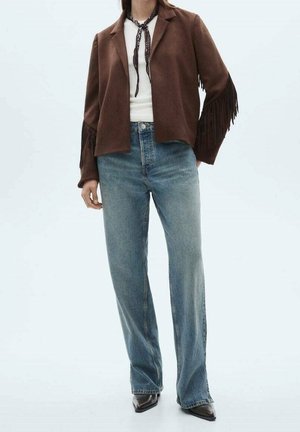 Femme portant une veste marron à franges, un haut blanc, un jean bleu droit et des chaussures noires à bout pointu, debout devant un fond uni.