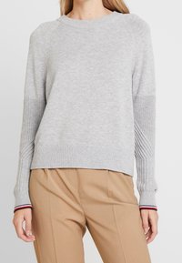 Woman wearing a light gray knit sweater with textured sleeves and striped cuffs, paired with high-waisted beige trousers.