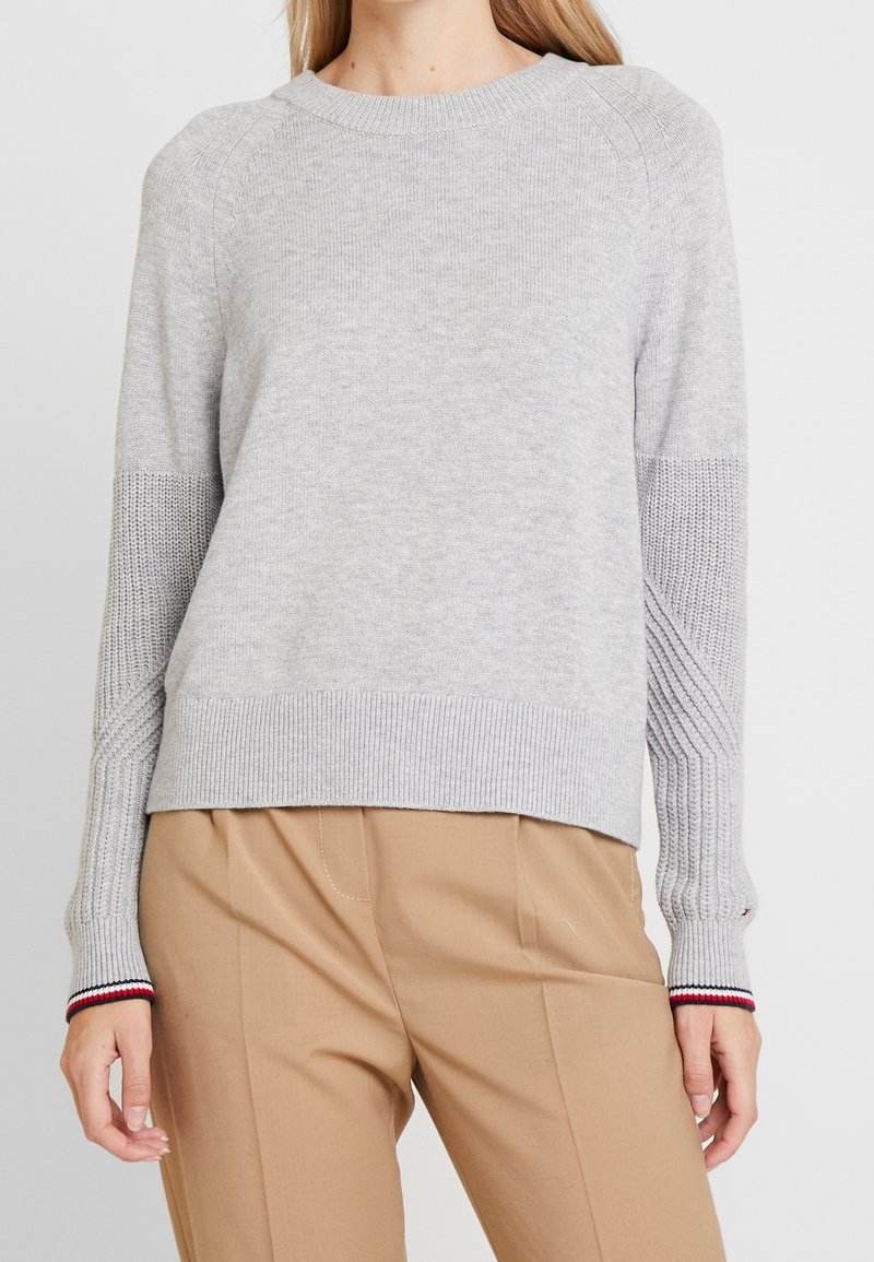 Woman wearing a light gray knit sweater with textured sleeves and striped cuffs, paired with high-waisted beige trousers.