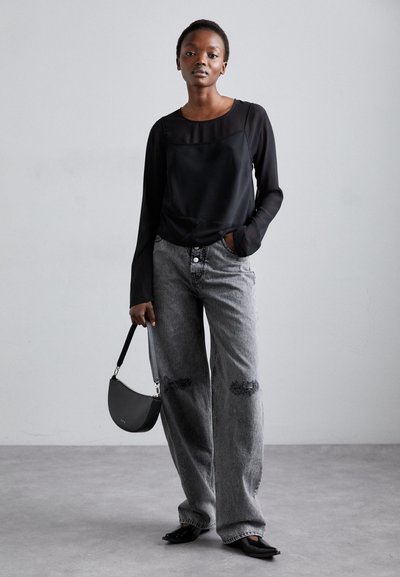 Blusa nera trasparente a maniche lunghe abbinata a jeans di denim grigi larghi con toppe strappate. Gli accessori includono una borsa curva nera.