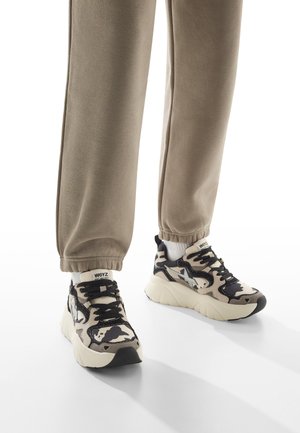 Le sneakers presentano un motivo camouflage beige e nero, materiali in mesh e suede, una suola bianca spessa e lacci neri. Abbinati a pantaloni della tuta khaki.