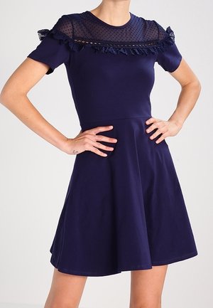 Laivastonsininen lyhythihainen mekko, jossa läpikuultava pilkkukuvioinen yläosa ja röyhelöyksityiskohta rinnan kohdalla, istuva vyötärö ja levenevä helma.