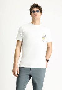 Camiseta blanca de manga corta con un diseño de bolsillo que presenta un patrón gráfico. Llevada con pantalones grises, el modelo posa frente a un fondo neutro.