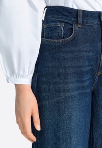 Tmavomodré rifľové nohavice s vysokým pásom, s ozdobným prešitím, predným vreckom a hladkou textúrou. Makro záber na oblasť pásu.