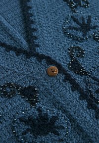 Blå stickad cardigan med texturerad tyg, med pärl- och broderidetaljer. En brun knapp syns nära mitten.