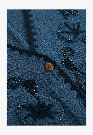 Lähikuva tummansinisestä teksturoidusta kankaasta, jossa on yksi ruskea nappi ja mustat helmet, jotka muodostavat kukkakuvion.