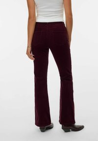 Mujer vestida con pantalones acampanados de pana de color púrpura oscuro, top sin mangas blanco y botas puntiagudas marrón oscuro, de pie contra un fondo sencillo.