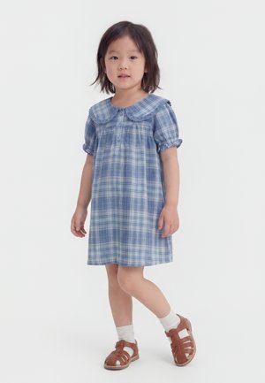 Νεαρό κορίτσι στέκεται, φοράει ένα μπλε καρό φόρεμα με φαρδύ γιακά, άσπρες κάλτσες και καφέ σανδάλια μπροστά σε απλό λευκό φόντο.
