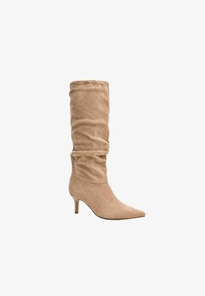 Høj beige ruskindsstøvle med spids tå, blød skaft og slank mellemhøj hæl, vist mod en hvid baggrund.