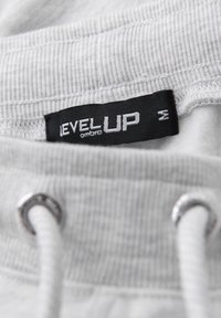 Gros plan de la ceinture d'un vêtement blanc avec une étiquette noire "LEVEL UP" taille M et des cordons blancs passés dans des œillets métalliques.