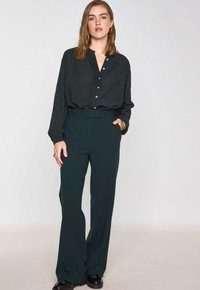 Lange mouwen, diep groene blouse met knopen en ruches, gecombineerd met een wijde broek in hetzelfde diep groen, beide gemaakt van een gladde stof.