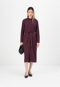 Mauve button-up dress with long sleeves, tie waist, and straight silhouette, paired with a black handbag and pointed-toe black heels.