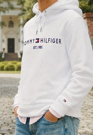 Person iført en hvid Tommy Hilfiger-hoodie med logo og "EST. 1985," hænderne i lommerne, stående udendørs på en brostensbelagt sti.