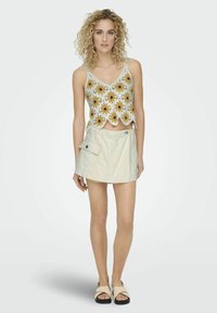 Gehaakt crop top met een geel en zwart bloemenpatroon, gecombineerd met een lichtbeige rok met een zijzak en knoopsluiting. Platte sandalen.