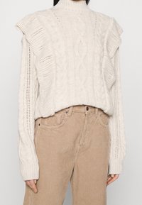 Pull en maille torsadée crème avec détails volantés aux épaules, porté avec un pantalon en velours côtelé beige taille haute.