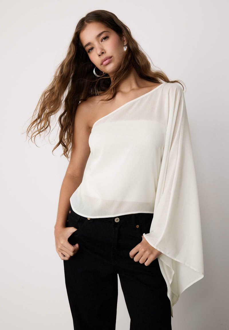 Jeune femme aux longs cheveux portant un top blanc à une épaule avec une manche fluide et un pantalon noir, les mains dans les poches, debout devant un fond neutre.