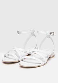 Hvide flade sandaler med flere krydsende stropper, ankelspænde og en lille hæl. Fremstillet af glat materiale med et minimalistisk design.