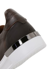 Gros plan sur le talon d'une basket en cuir marron foncé avec une bande métallique au-dessus d'une épaisse semelle blanche sur fond blanc.