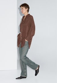 Brun, tekstureret skjorte med knapper og sidelommer, kombineret med lyseblå jeans og sorte spidse sko, stående mod en neutral baggrund.