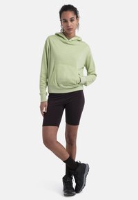 Sudadera con capucha de color verde claro y bolsillo frontal, combinada con pantalones cortos de ciclismo marrón oscuro y zapatos de senderismo negros. Textura suave, corte moderno.