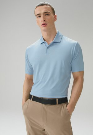 Young man with short hair wearing a light blue short-sleeve polo shirt and beige pants with a black belt, standing with hands in pockets.