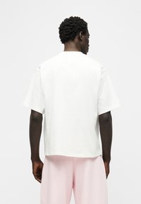 Άνδρας με κοντά σγουρά μαλλιά, γυρισμένος από την πλάτη, φοράει ένα απλό λευκό κοντομάνικο πουκάμισο και ανοιχτό ροζ παντελόνι, σε λευκό φόντο.