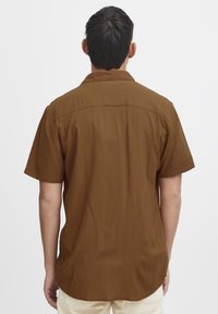 Hombre con pelo oscuro corto, vistiendo una camisa marrón de manga corta y pantalones de color claro, de espaldas contra un fondo blanco liso.