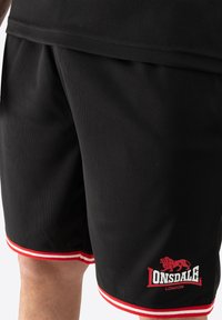 Čierne atletické šortky z priedušnej tkaniny s červenými a bielymi pruhmi a výrazným logom Lonsdale London na dolnej časti nohavice.