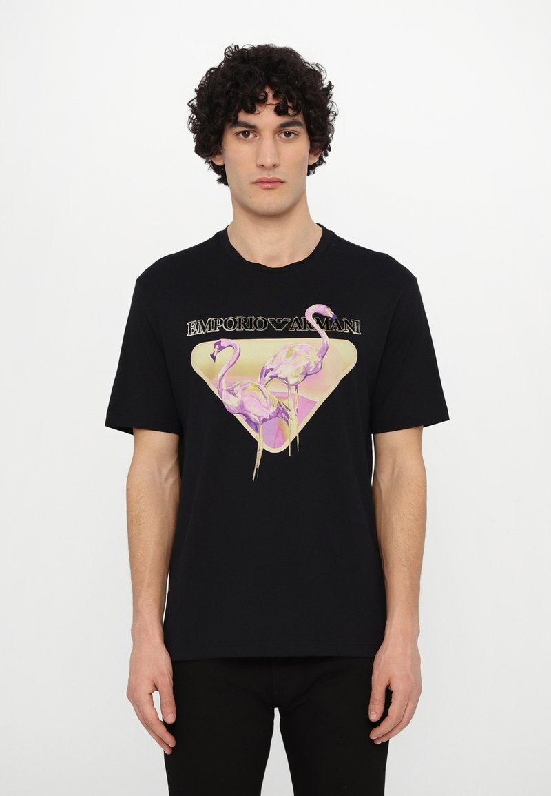 Črna bombažna majica s kratkimi rokavi, ki prikazuje trikotno grafiko z dvema rožnatima flamingoma in napisom "EMPORIO ARMANI" v zlati barvi, z okroglim izrezom.