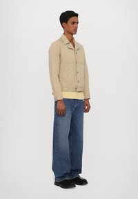 Giacca beige con dettagli in rivetti argentati, chiusura a bottone e vestibilità rilassata. Abbinata a jeans blu larghi e scarpe nere.