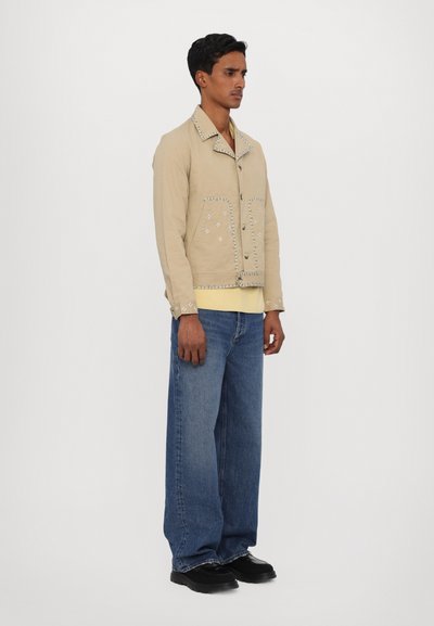 Beige jacket with silver stud detailing, button closure, and a relaxed fit. Paired with baggy blue jeans and black shoes.