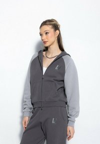 Młoda kobieta ubrana w szaro-czarną bluzę z kapturem na zamek i dopasowane dresy z logo, stojąca na tle jasnej białej ściany.