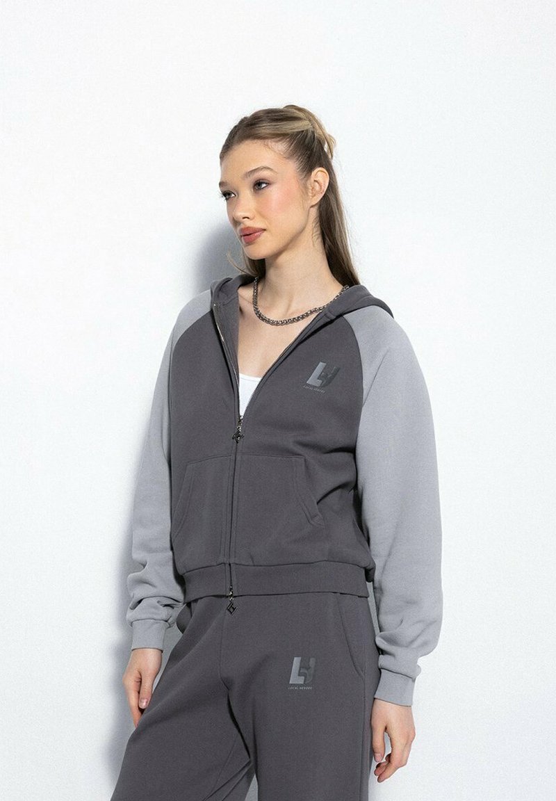Młoda kobieta ubrana w szaro-czarną bluzę z kapturem na zamek i dopasowane dresy z logo, stojąca na tle jasnej białej ściany.