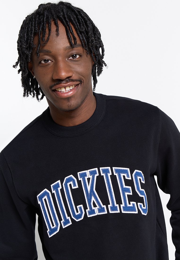 Uomo sorridente con dreadlocks corti indossa una felpa nera con grande scritta blu e bianca "DICKIES" sul davanti su sfondo bianco.