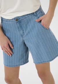 Blå denimshorts har vita vertikala ränder, en framficka och en knäppning i midjan. Tyget verkar mjukt och hållbart.