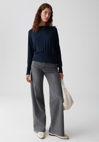 Marineblaues Langarmshirt mit geraffter Taille, kombiniert mit grauen weit geschnittenen Jeans und beigen Schuhen, dazu eine kleine cremefarbene Handtasche.
