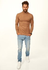 Brauner Rollkragenpullover aus gerippter Stoff, kombiniert mit hellblauen Jeans und weißen Turnschuhen. Schlichte Gestaltung mit körperbetonter Silhouette.
