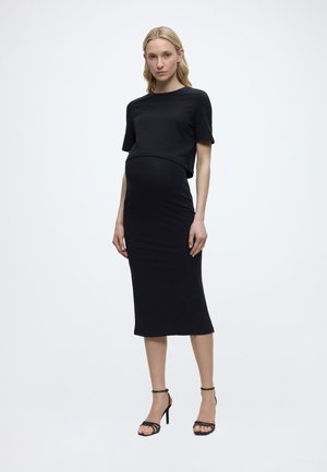 Terhes nő, aki egyenesen áll, fekete rövid ujjú felsőt és fekete, testhez simuló midi szoknyát visel, fekete magas sarkú szandálban.