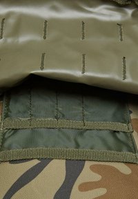 Kamuflāžas auduma soma ar gludu olīvu zaļu virsmu, kas iezīmēta ar vertikālām šuvēm un teksturētu olīvu zaļu jostu.